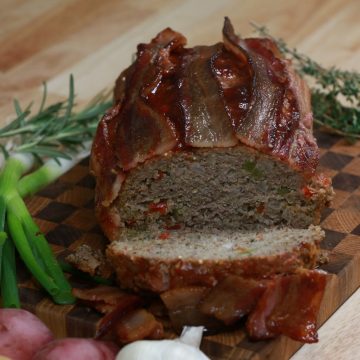Sliced gluten-free BBQ meatloaf topped with bacon, showing quinoa and diced peppers inside, on a wooden board with thyme, green onions and potatoes