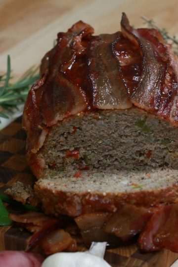 Sliced gluten-free BBQ meatloaf topped with bacon, showing quinoa and diced peppers inside, on a wooden board with thyme, green onions and potatoes