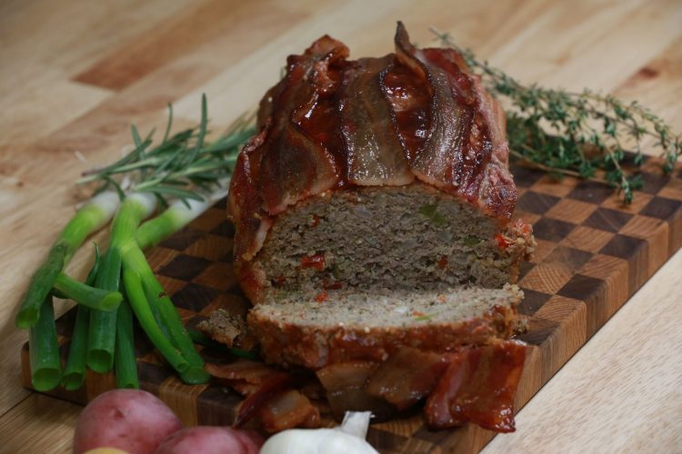 Sliced gluten-free BBQ meatloaf topped with bacon, showing quinoa and diced peppers inside, on a wooden board with thyme, green onions and potatoes