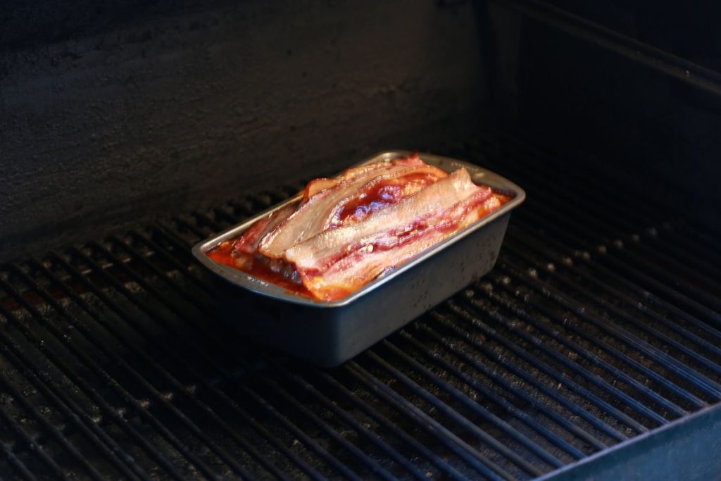 Bacon-topped BBQ meatloaf in a loaf pan cooking in a smoker