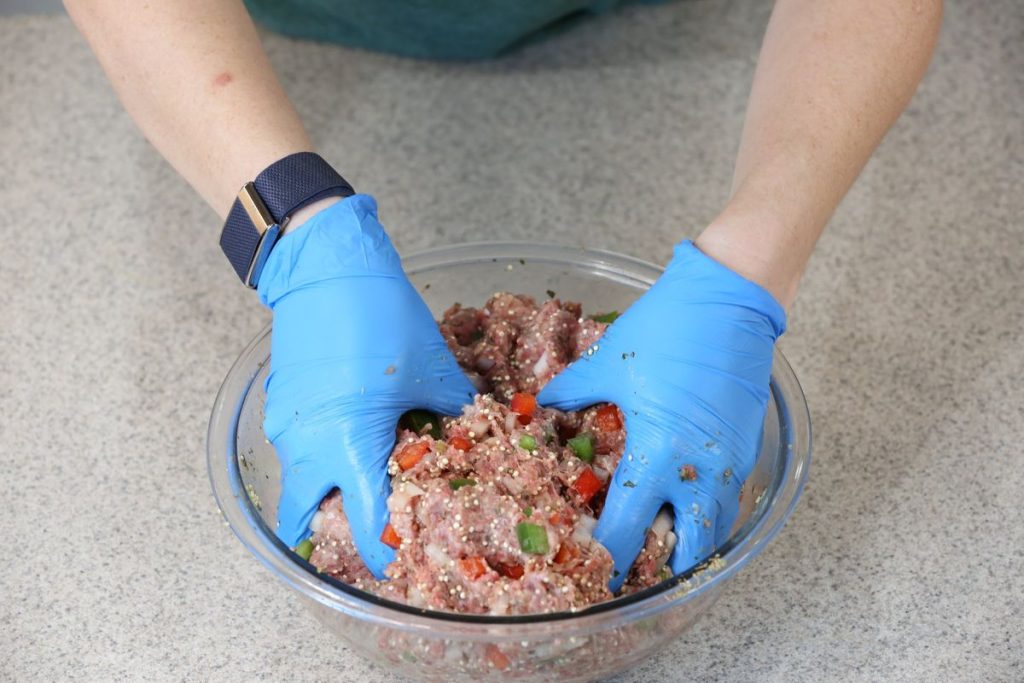 Gloved hands mixing ground meat, diced peppers and dry quinoa in a glass bowl